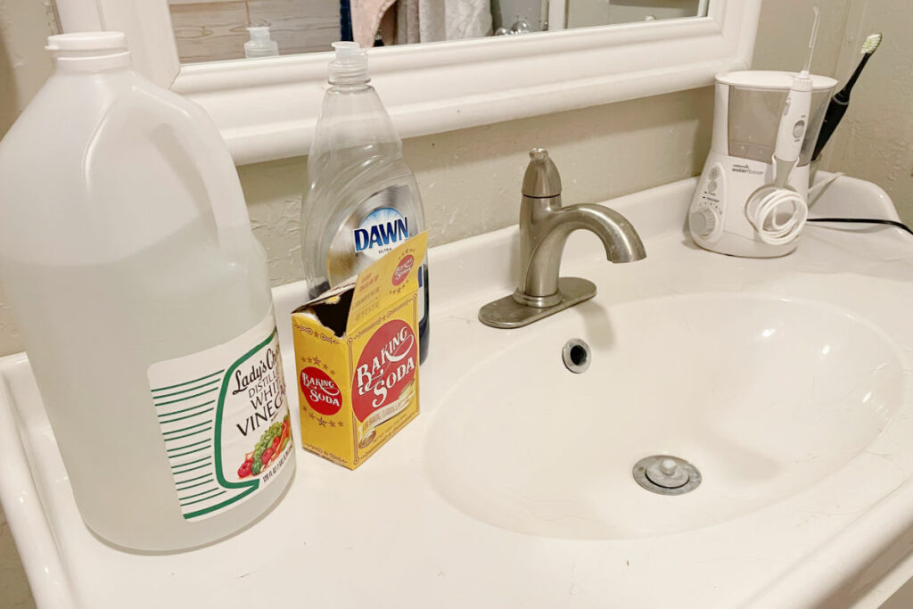 Vinegar, baking soda and Dawn dish soap placed on a dirty white porcelain sink. 
