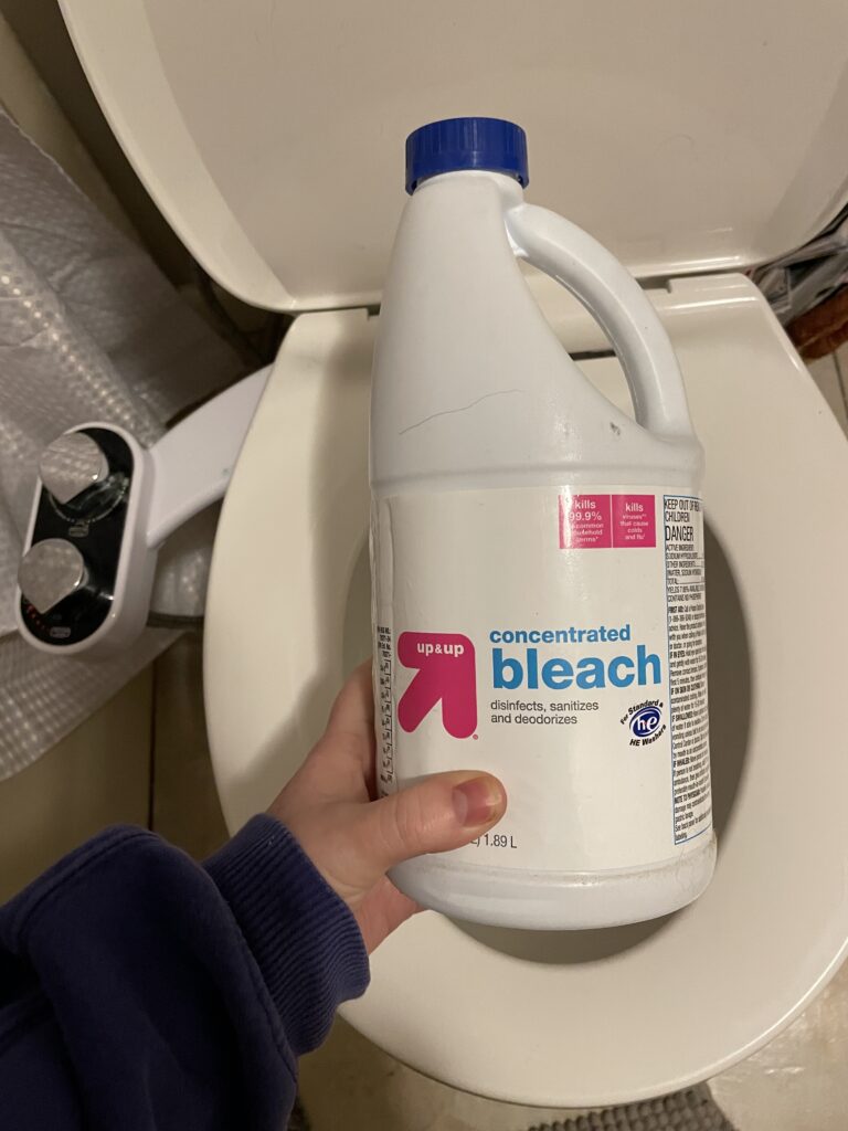 A bottle of bleach held in front of a toilet bowl that will be used to clean a plunger. 