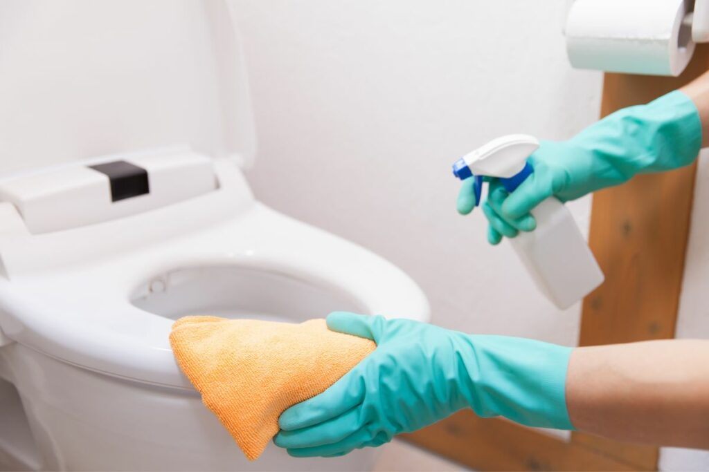 Close up of hands wearing gloves cleaning a toilet seat with a spray bottle and rag. 