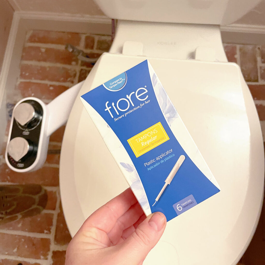 A box of tampons held in front of a toilet. 